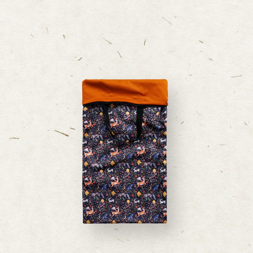 A tall rectangular Eco Mini brand wet bag with black handles, featuring a dense all-over pattern of stylized forest animals in purple, orange, and white on a dark navy background, set against a textured off-white paper backdrop.