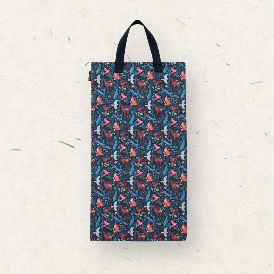 A cloth diaper wet bag with a dark navy blue background and an all-over whimsical pattern of red foxes, pink fox tails, and white birds. The bag features two black fabric handles and is photographed hanging against a textured off-white paper background.