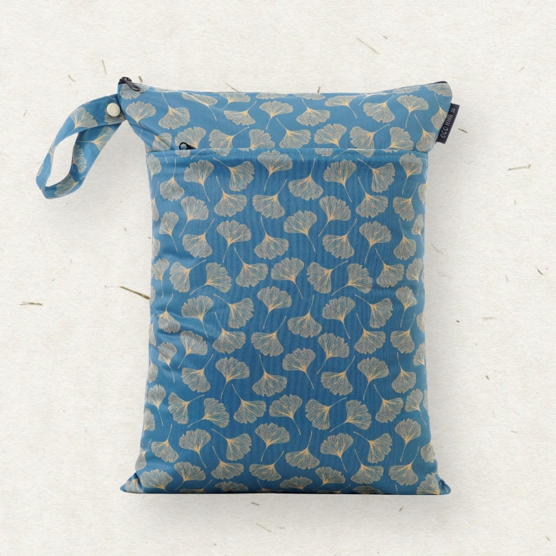 A blue wet bag with a beige ginkgo leaf pattern, featuring a matching fabric loop strap and a small black brand tag on the right side, laid flat on a textured off-white background.