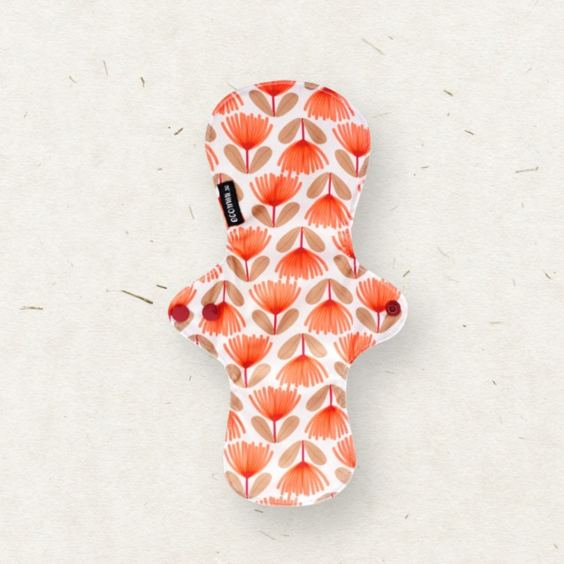 A reusable menstrual pad with a white background and a repeating pattern of orange and beige palm leaf motifs, shown on a textured off-white surface. A small black fabric tag with the brand name 'Eco Mini' is visible on the left side.