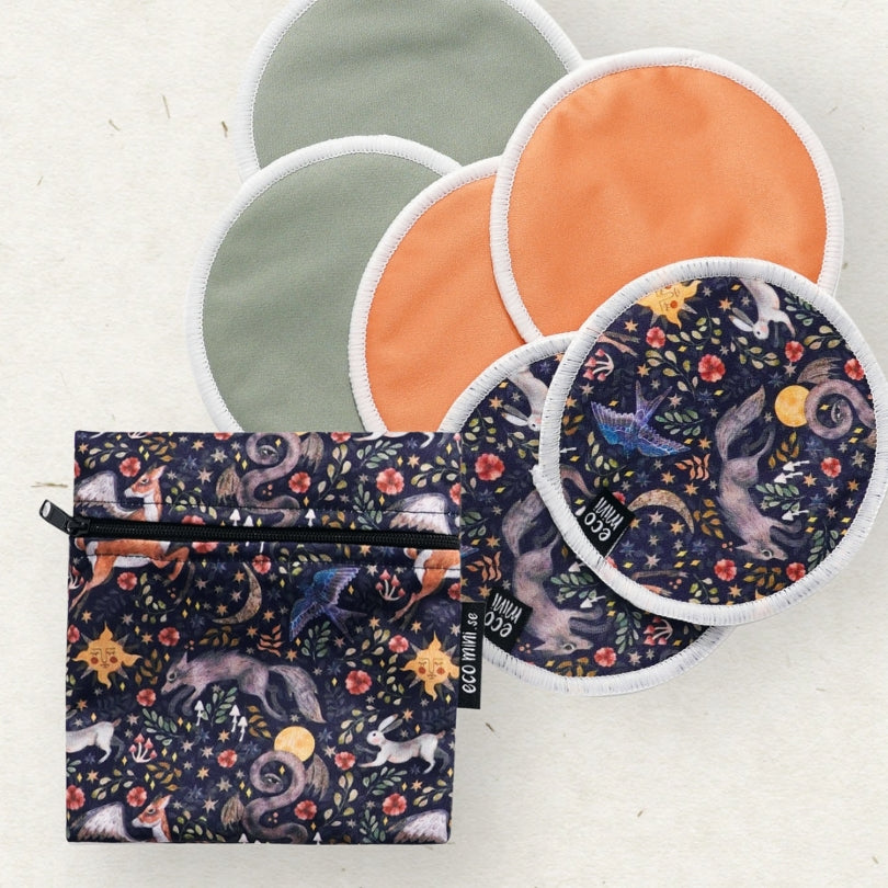 A set of Eco Mini reusable cloth breastfeeding pads with white trim and a matching zippered pouch, both featuring a whimsical dark blue animal and nature print. The pads are shown in various colors, arranged on a light beige textured background.