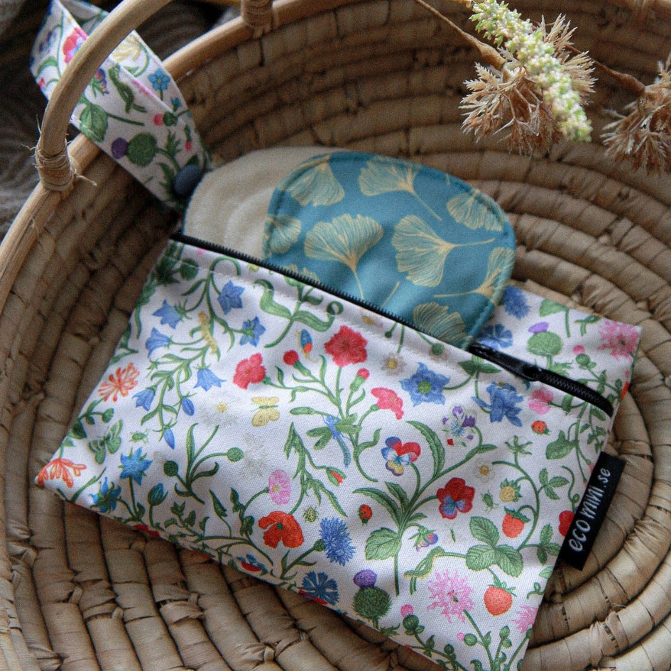 A fabric wet bag with a colorful wildflower print on a white background, placed inside a woven wicker basket next to a teal pouch with a ginkgo leaf pattern. A black fabric tag on the pouch reads 'eco mini'.