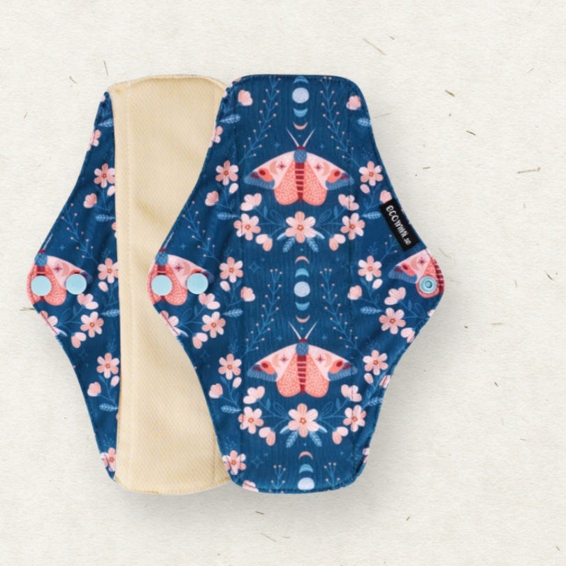 A reusable cloth menstrual pad with a navy blue background, pink and orange moths, and pink flowers, shown with its beige lining on a light textured background.