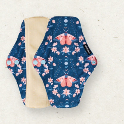 A reusable cloth menstrual pad with a navy blue background, pink and orange moths, and pink flowers, shown with its beige lining on a light textured background.