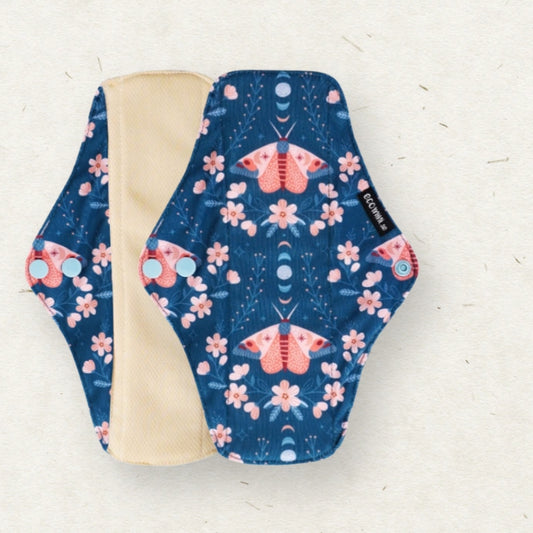 A reusable cloth menstrual pad with a navy blue background, pink and orange moths, and pink flowers, shown with its beige lining on a light textured background.