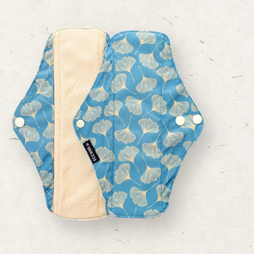 A reusable cloth menstrual pad shown flat on a light textured background. The pad has a blue exterior with a white ginkgo leaf pattern, a beige inner lining, and white snap closures.