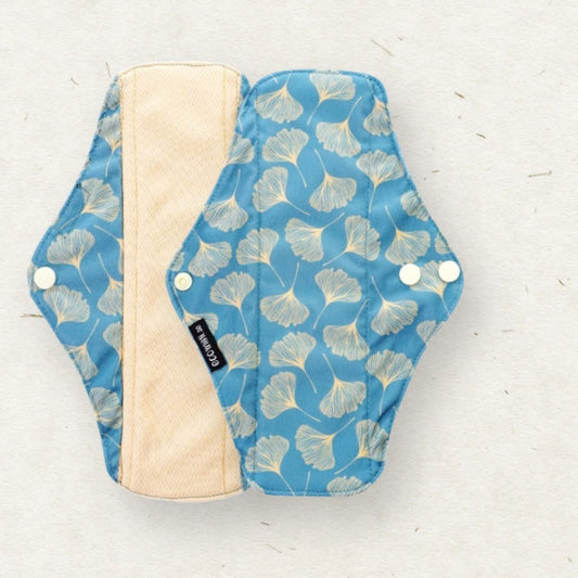 A reusable cloth menstrual pad shown flat on a light textured background. The pad has a blue exterior with a white ginkgo leaf pattern, a beige inner lining, and white snap closures.