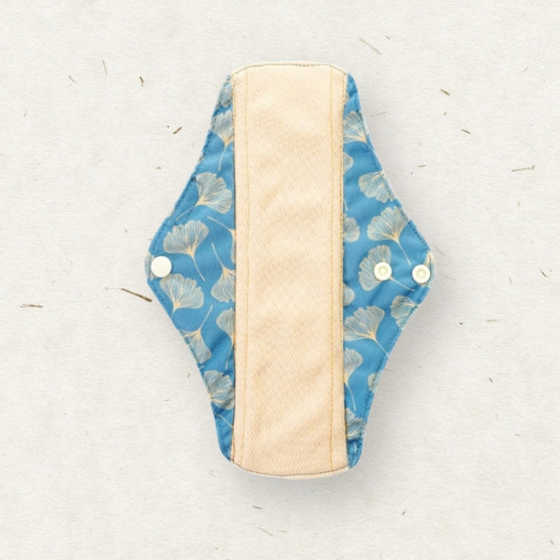 A reusable cloth menstrual pad with a blue ginkgo leaf pattern on the outer fabric and a beige textured center lining, shown on a light textured background.