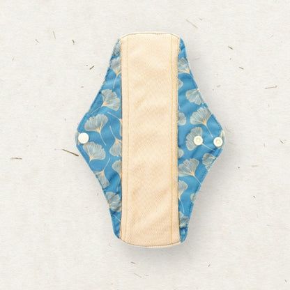 A reusable cloth menstrual pad with a blue ginkgo leaf pattern on the outer fabric and a beige textured center lining, shown on a light textured background.