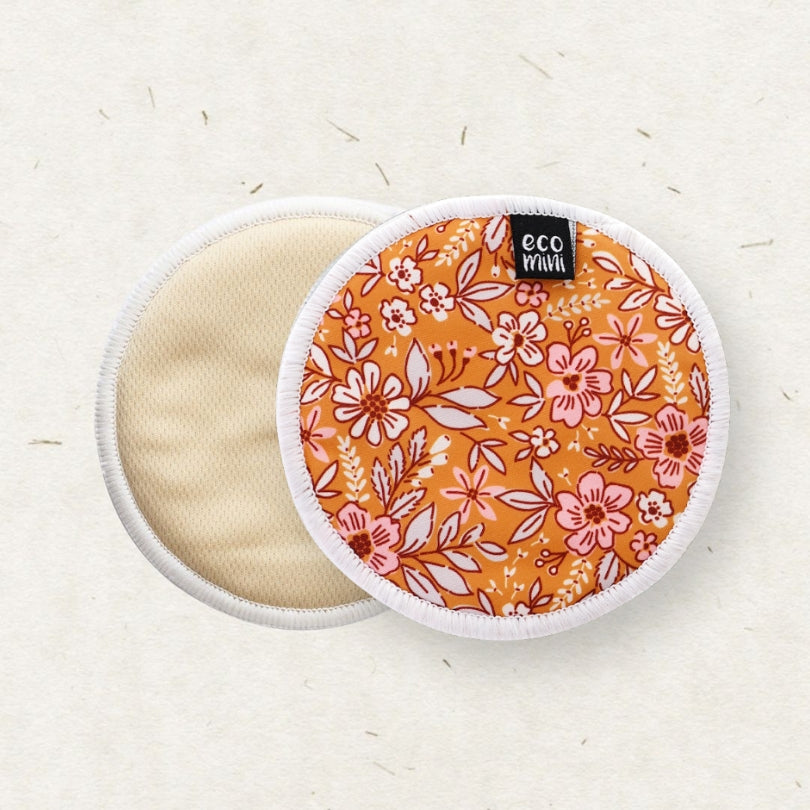 Two reusable Eco Mini Breast pads on a light beige textured background. One pad shows a warm orange floral pattern with pink and white flowers, while the other shows a plain beige fabric side with a white stitched edge.