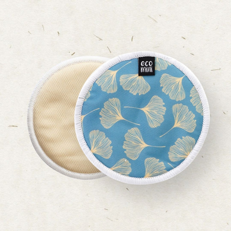Two round reusable cloth breast pads by ECO MINI, showing the blue side with a gold ginkgo leaf pattern and the beige fabric side, on a textured off-white background.