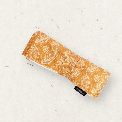 Eco Mini Changing mat/ Skötunderlägg - with an orange and white mandala pattern, placed diagonally on a textured off-white background with small scattered fibers.