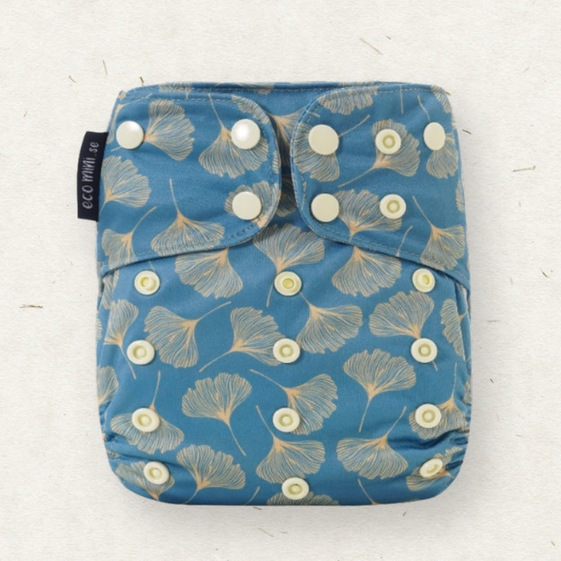 A reusable cloth diaper with a blue background and a beige ginkgo leaf pattern, featuring white snap closures and a black brand tag reading 'eco mini'.