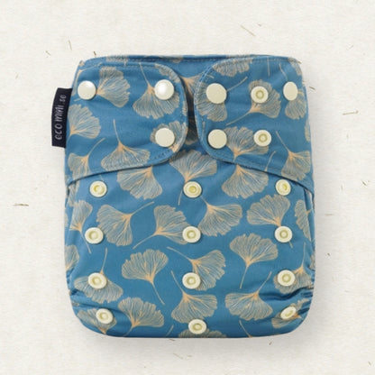 A reusable cloth diaper with a blue background and a beige ginkgo leaf pattern, featuring white snap closures and a black brand tag reading 'eco mini'.