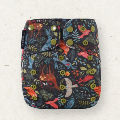 A reusable cloth diaper with a dark background and a colorful nature print featuring birds, leaves, and flowers, secured with three metal snaps. A black fabric tag on the left side reads 'ECO MINI'.