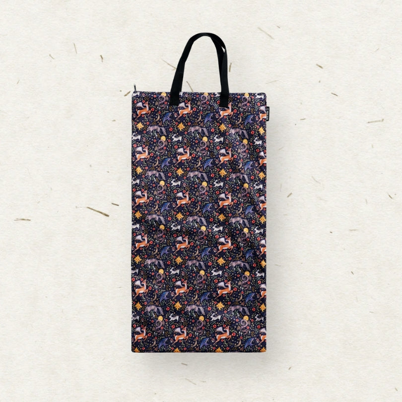 A tall rectangular Eco Mini brand wet bag with black handles, featuring a dense all-over pattern of stylized forest animals in purple, orange, and white on a dark navy background, set against a textured off-white paper backdrop.