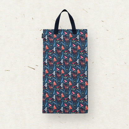 A cloth diaper wet bag with a dark navy blue background and an all-over whimsical pattern of red foxes, pink fox tails, and white birds. The bag features two black fabric handles and is photographed hanging against a textured off-white paper background.