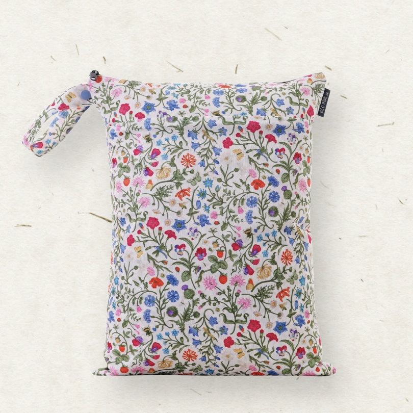 A cloth diaper wet bag with a colorful wildflower print on a white background, featuring a top zipper and a matching wrist strap, photographed on a textured off-white surface.