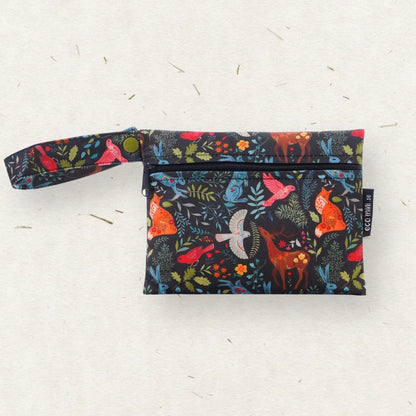 A small wet bag with a wrist strap, featuring a colorful woodland print of birds, foxes, and leaves on a black background, photographed on a light textured surface.