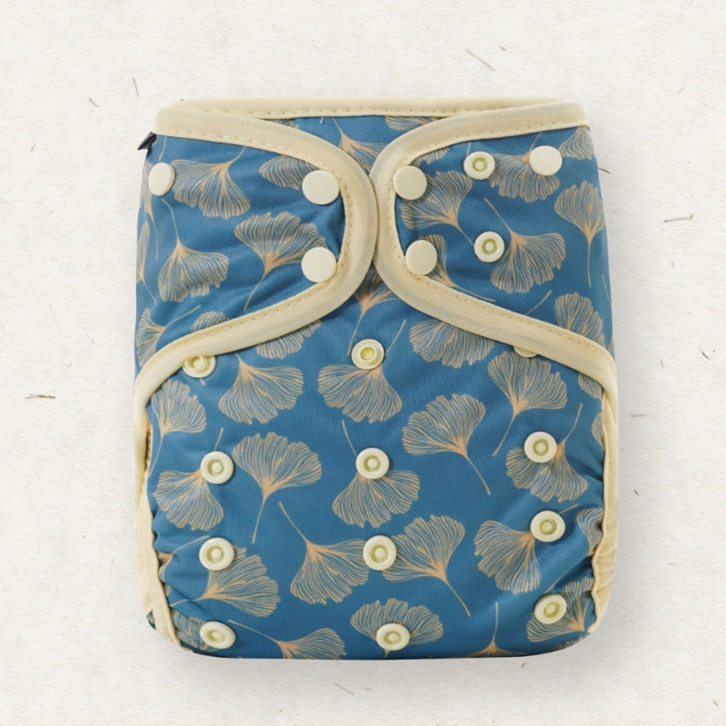 A reusable cloth diaper with a blue background and a beige ginkgo leaf pattern, featuring adjustable snap closures and beige fabric trim, laid flat on a light textured surface.