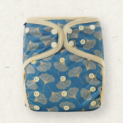 A reusable cloth diaper with a blue background and a beige ginkgo leaf pattern, featuring adjustable snap closures and beige fabric trim, laid flat on a light textured surface.