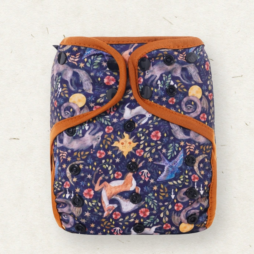 A reusable cloth diaper with a dark navy blue background printed with a whimsical celestial and animal pattern, featuring orange-brown trim and black snap closures, shown on a light textured background.