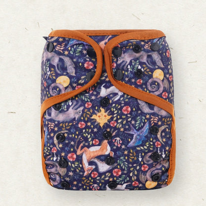 A reusable cloth diaper with a dark navy blue background printed with a whimsical celestial and animal pattern, featuring orange-brown trim and black snap closures, shown on a light textured background.