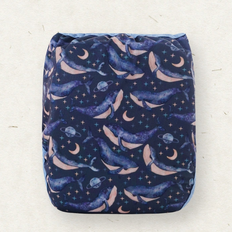 A reusable cloth diaper with a space-themed print of whales, moons, stars, and planets on a dark navy blue background, shown flat against a light beige textured background.