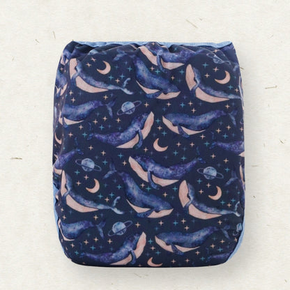 A reusable cloth diaper with a space-themed print of whales, moons, stars, and planets on a dark navy blue background, shown flat against a light beige textured background.