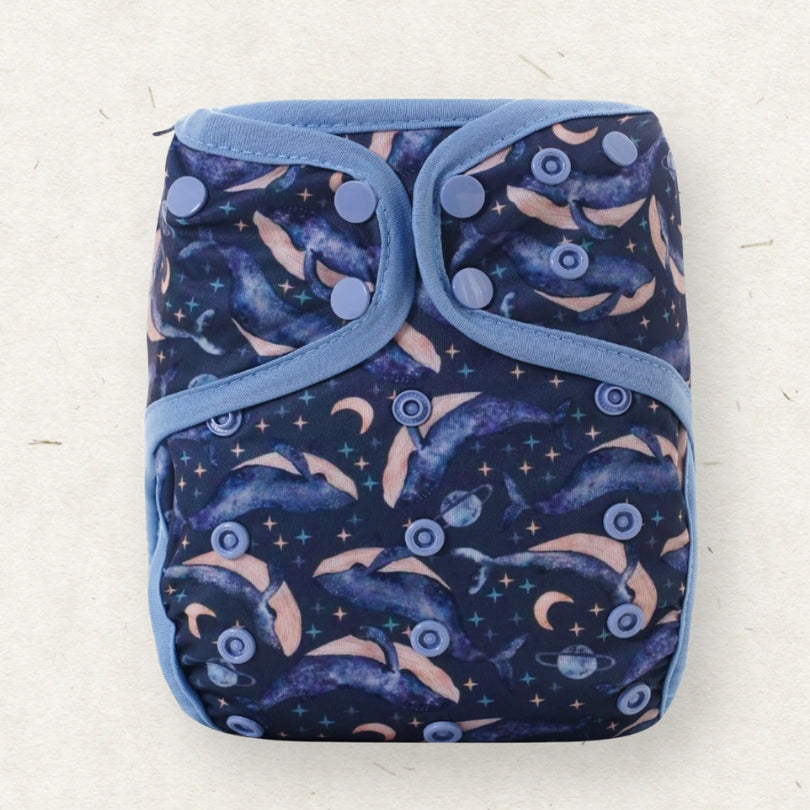 A reusable cloth diaper with a dark navy blue background featuring a pattern of dolphins, crescent moons, stars, and planets, bordered by light blue trim and secured with snap buttons.