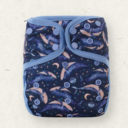 A reusable cloth diaper with a dark navy blue background featuring a pattern of dolphins, crescent moons, stars, and planets, bordered by light blue trim and secured with snap buttons.