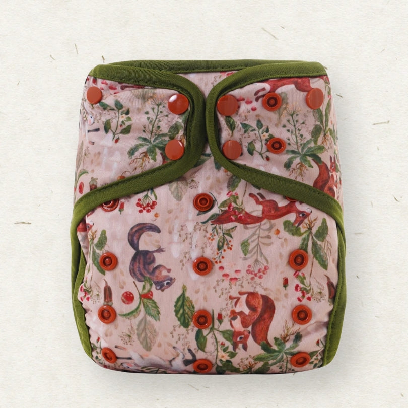 A reusable cloth diaper with a woodland squirrel pattern on a light pink background, featuring olive green trim and red snap closures, photographed flat on a textured off-white surface.