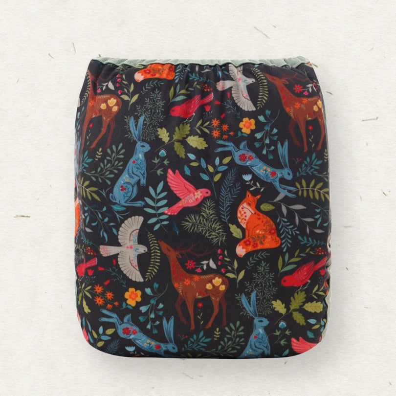A square reusable cloth diaper with a dark navy background and a colorful woodland animal print featuring blue rabbits, red birds, orange foxes, and green leaves, shown against a light off-white textured background.