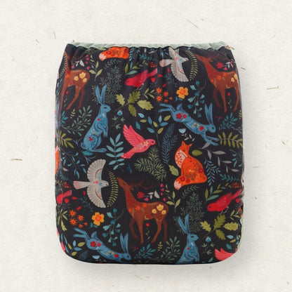 A square reusable cloth diaper with a dark navy background and a colorful woodland animal print featuring blue rabbits, red birds, orange foxes, and green leaves, shown against a light off-white textured background.