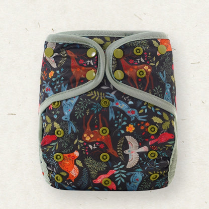 A reusable cloth diaper with a dark navy background and a colorful woodland print featuring deer, birds, flowers, and leaves. It has sage green trim and olive green snap closures, laid flat on a light textured background.