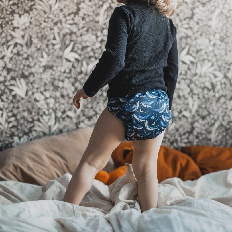 Child wearing blue patterned Eco mini cloth diaper standing on a bed with a textured wall in the background