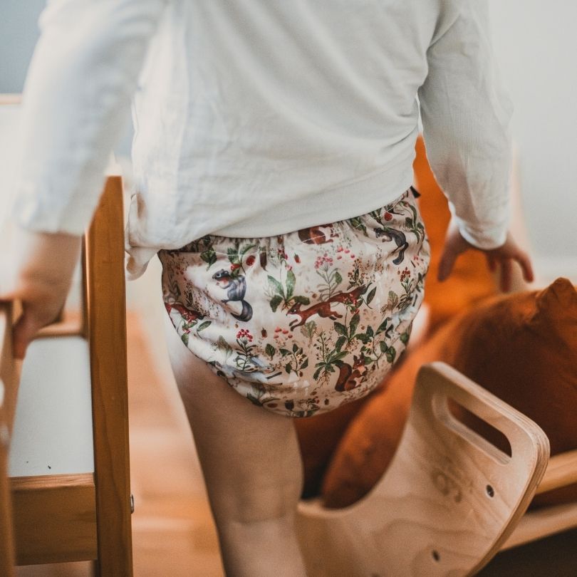 Person wearing a patterned Eco Mini cloth diaper standing near wooden furniture.