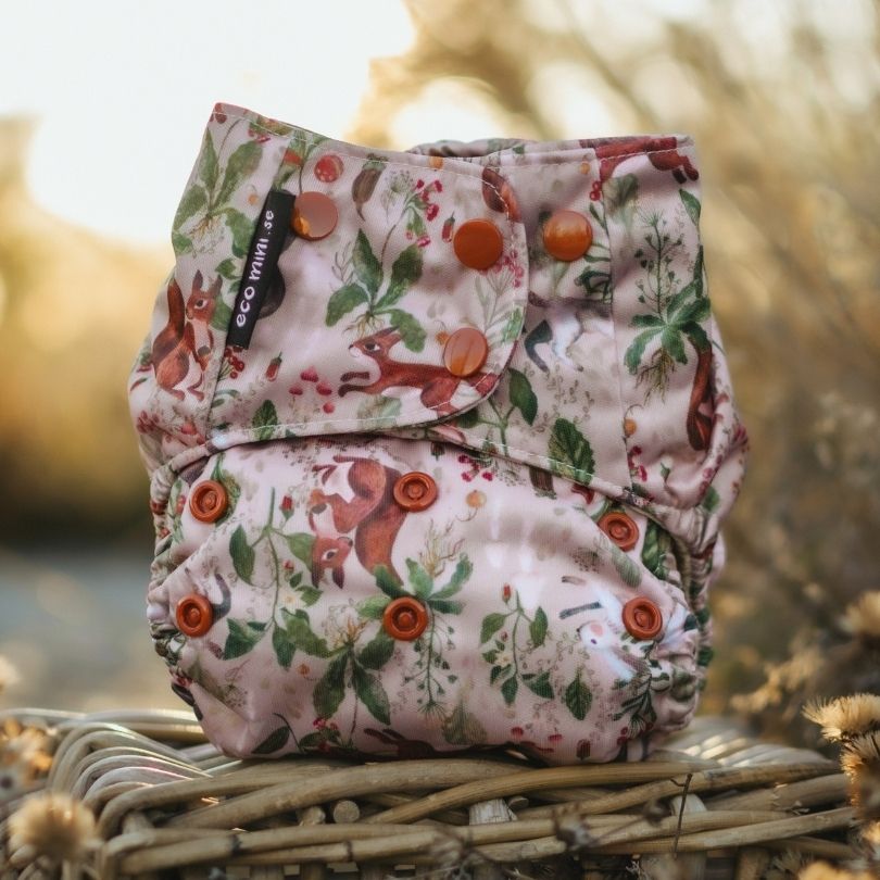 Eco Mini floral-patterned cloth diaper with orange buttons on a wooden surface with a blurred natural background
