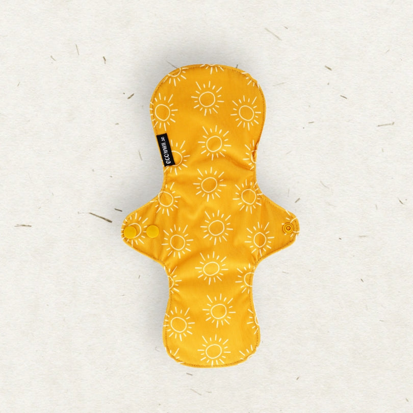 A yellow menstrual pad with a white sun pattern, laid flat on a textured off-white background. A small black brand tag reading 'Eco Mini' is visible on the left side.