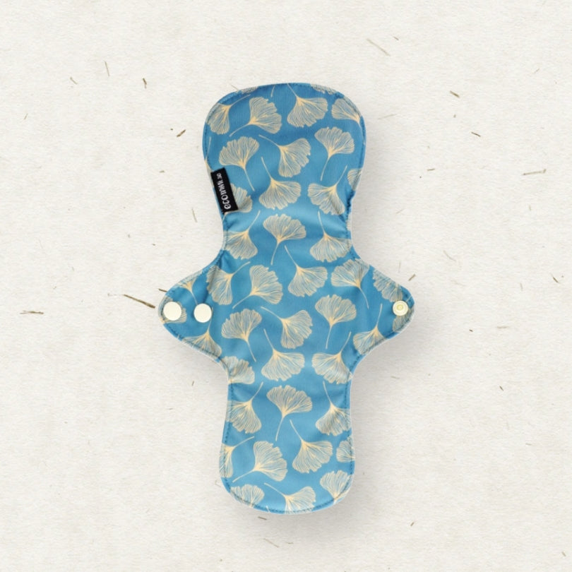 A reusable fabric menstrual pad with a blue background and a light beige ginkgo leaf pattern, laid flat on a textured off-white paper background. A small black brand tag reading 'Eco Mini' is visible on the left side.