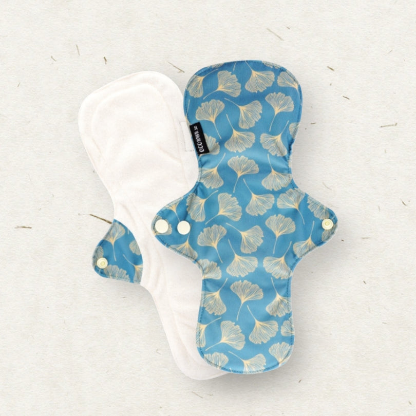 Two reusable cloth menstrual pads on a light textured background. One pad is shown from the patterned side, featuring a blue base with a light beige ginkgo leaf print, and the other is shown from the plain side in white.