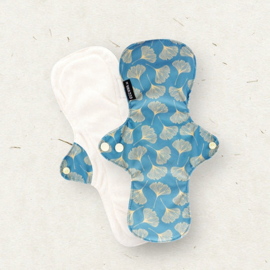 Two reusable cloth menstrual pads on a light textured background. One pad is shown from the patterned side, featuring a blue base with a light beige ginkgo leaf print, and the other is shown from the plain side in white.