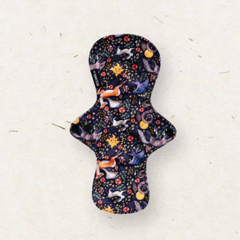 A reusable fabric menstrual pad with a dark navy blue background and a whimsical print of fairies, stars, and flowers, laid flat on a textured off-white surface.