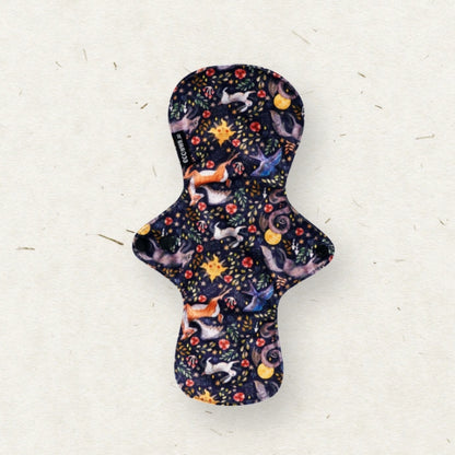 A reusable fabric menstrual pad with a dark navy blue background and a whimsical print of fairies, stars, and flowers, laid flat on a textured off-white surface.