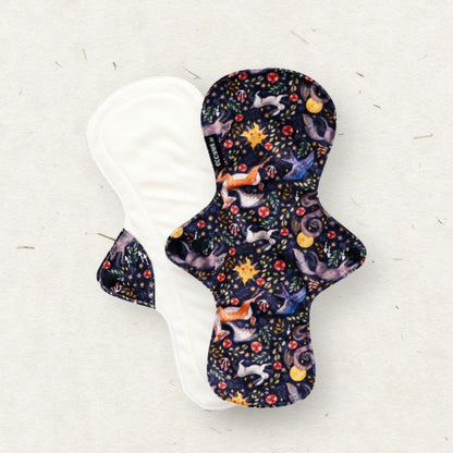 A reusable fabric menstrual pad shown from both sides on a light textured background. The front side features a dark navy blue background with a whimsical pattern of foxes, stars, and flowers in orange, yellow, and light blue.