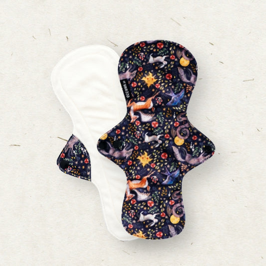 A reusable fabric menstrual pad shown from both sides on a light textured background. The front side features a dark navy blue background with a whimsical pattern of foxes, stars, and flowers in orange, yellow, and light blue.