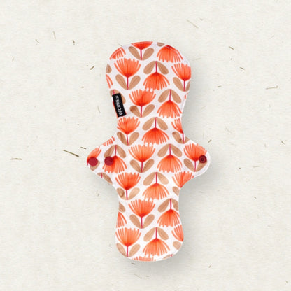 A reusable menstrual pad with a white background and a repeating pattern of orange and beige palm leaf motifs, shown on a textured off-white surface. A small black fabric tag with the brand name 'Eco Mini' is visible on the left side.