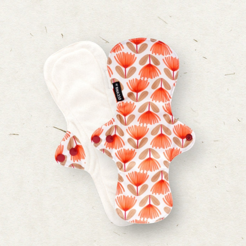 Two reusable cloth menstrual pads on a light textured background. One pad shows a white back with a small black brand tag, and the other shows the front with a white base and a repeating orange and beige floral pattern.