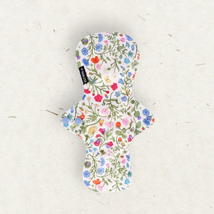 A single menstrual pad with a colorful wildflower print on a white background, laid flat on a textured off-white paper surface. A small black brand tag reading 'Eco Mini' is visible on the left side.