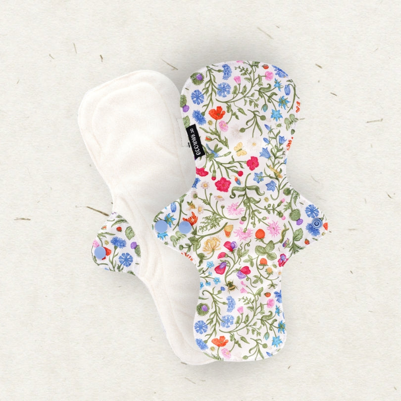 Two reusable cloth menstrual pads on a light textured background. One pad is shown from the back in solid white fabric, and the other is shown from the front with a dense multicolor floral print featuring green leaves and red, blue, pink, and yellow flowers on a white base.